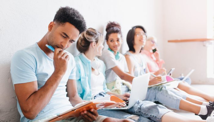 Students sitting on the floor with laptops and doing their homework together. Black boy with trendy hairstyle thinking about difficult task, holding pen in hand.